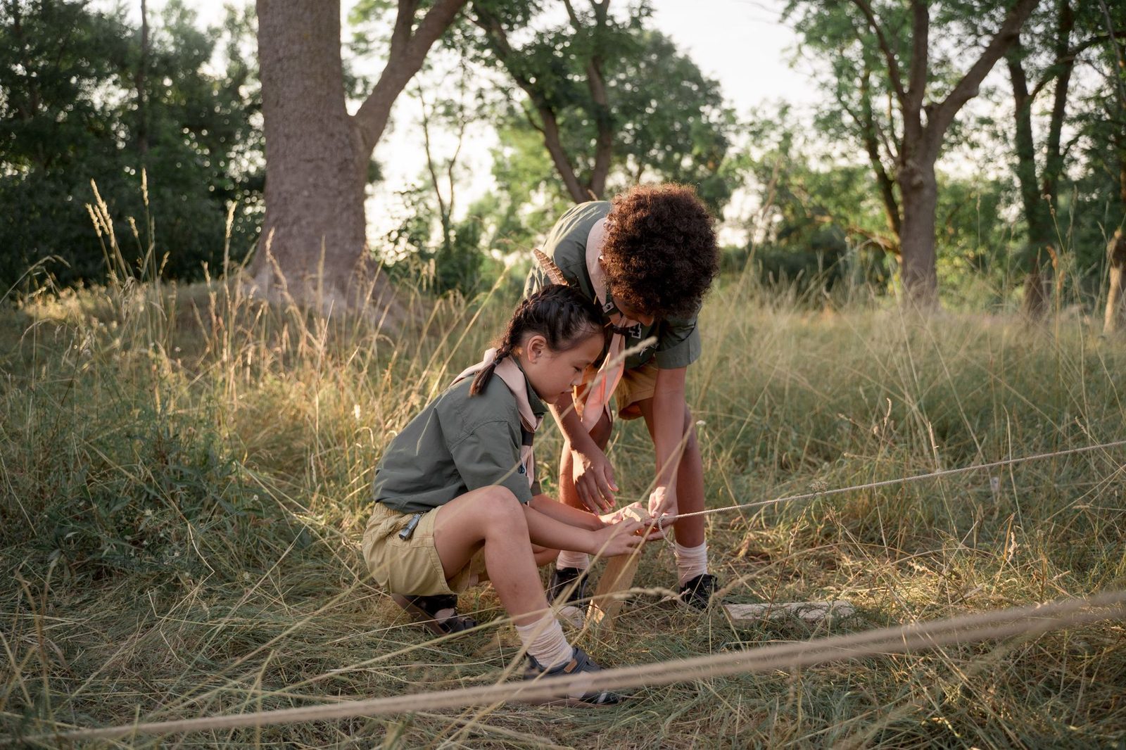 Two young scouts tying knots in a grassy campsite, focused on teamwork and learning.