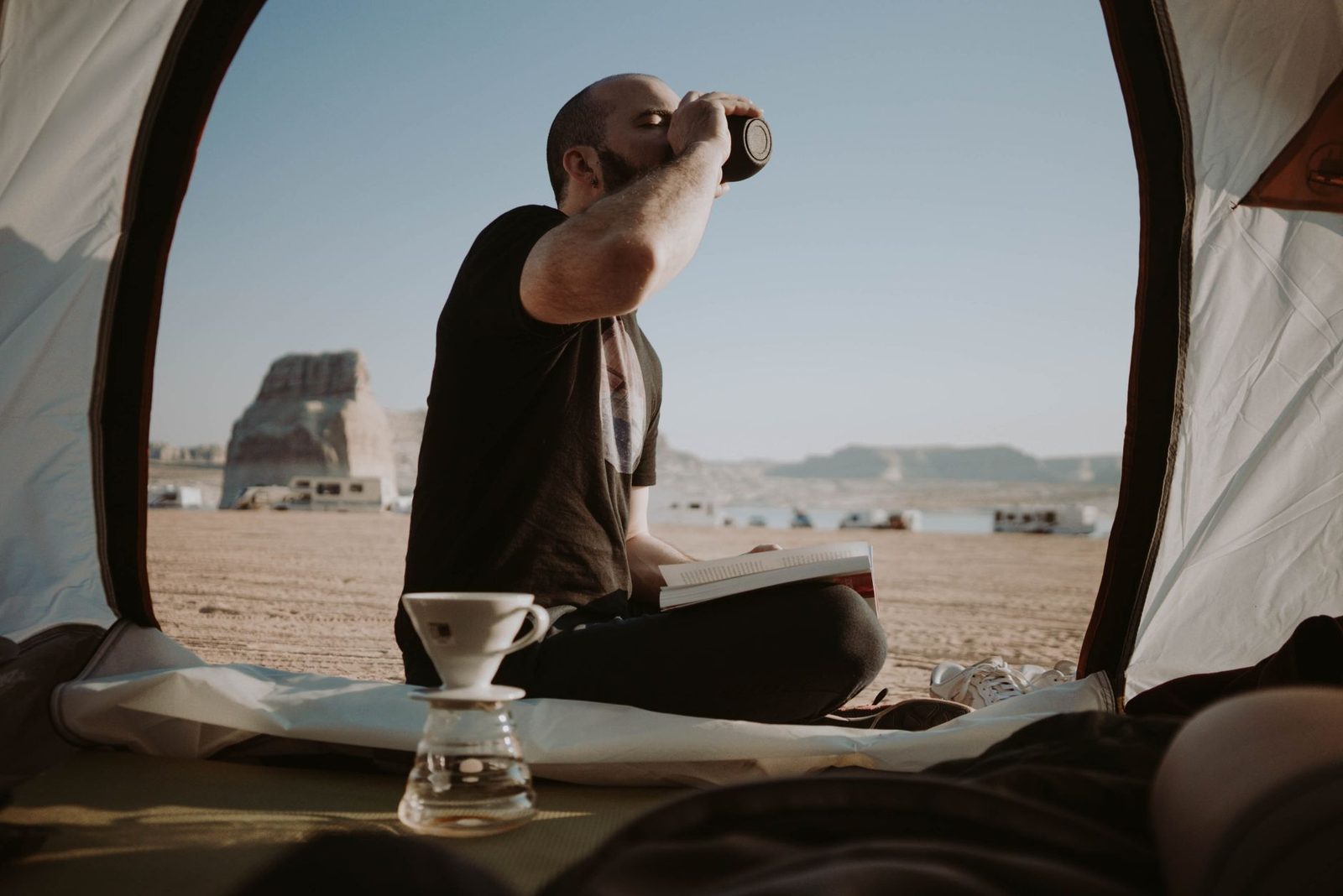 Side view of male traveler sitting in tent with book near pour over cup while drinking hot beverage in nature during trip