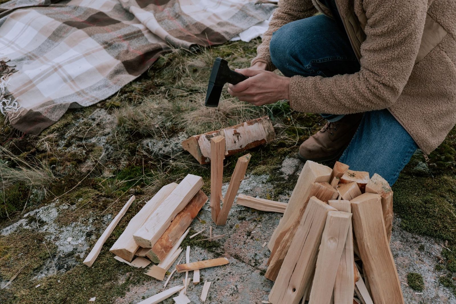 Person splits logs with an axe on grassy ground, surrounded by firewood and autumn blanket.