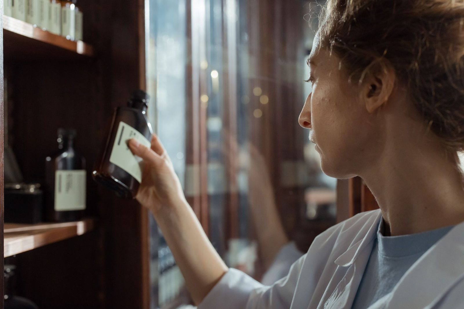 A pharmacist in a lab coat selecting a medication bottle from a pharmacy shelf indoors.