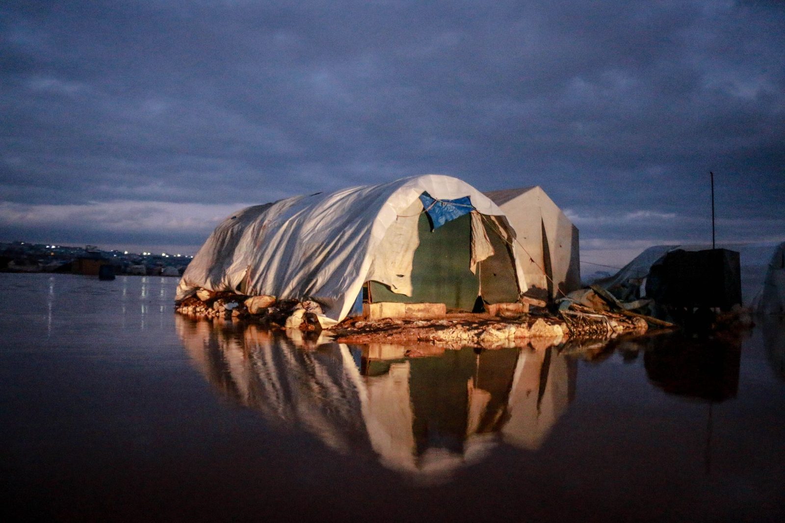 Poor tent with crumpled fabric reflecting in rippled lake against constructions under cloudy sky in evening