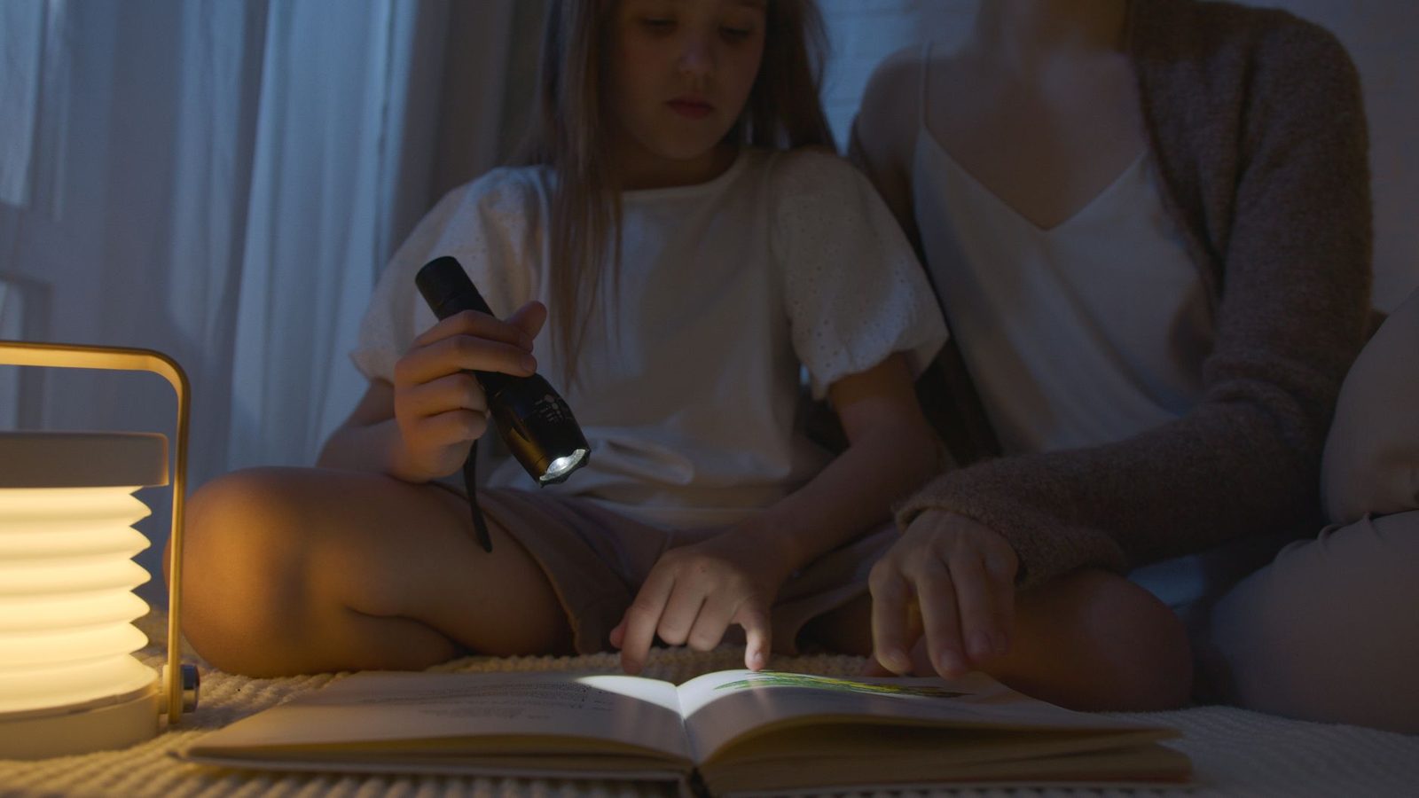A child and adult reading a book at night using a flashlight, creating a cozy atmosphere.