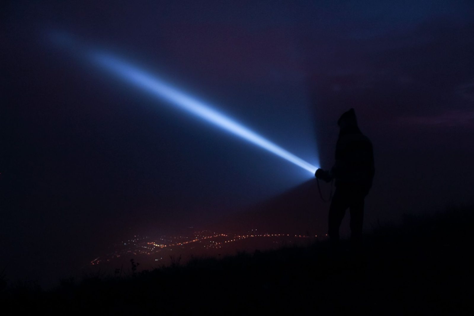 A person holds a flashlight, casting a beam at night with city lights in the background in Virgínia, Brazil.