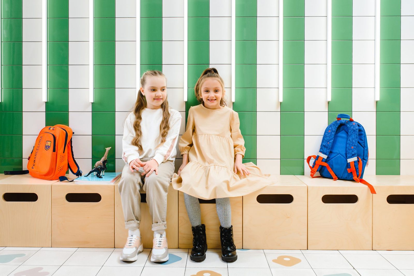 Two young girls sitting happily on a bench with backpacks inside a classroom.
