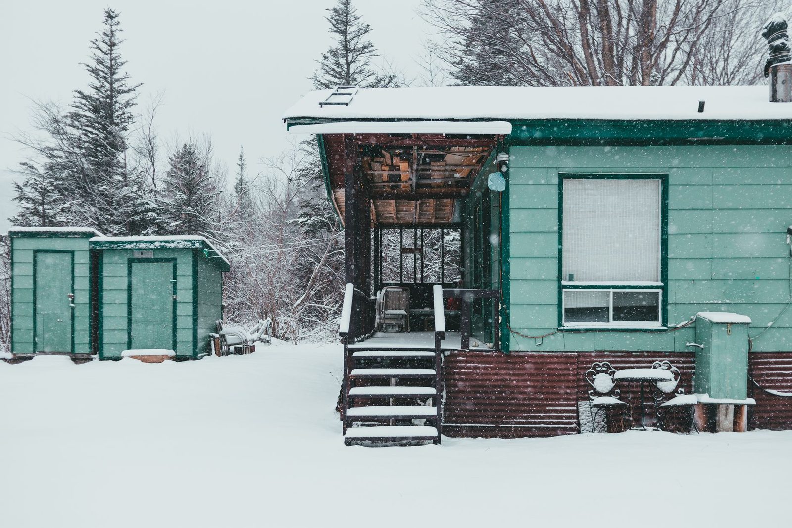 Wooden house near small buildings and snowy trees in forest in winter day in countryside under gray cloudless sky