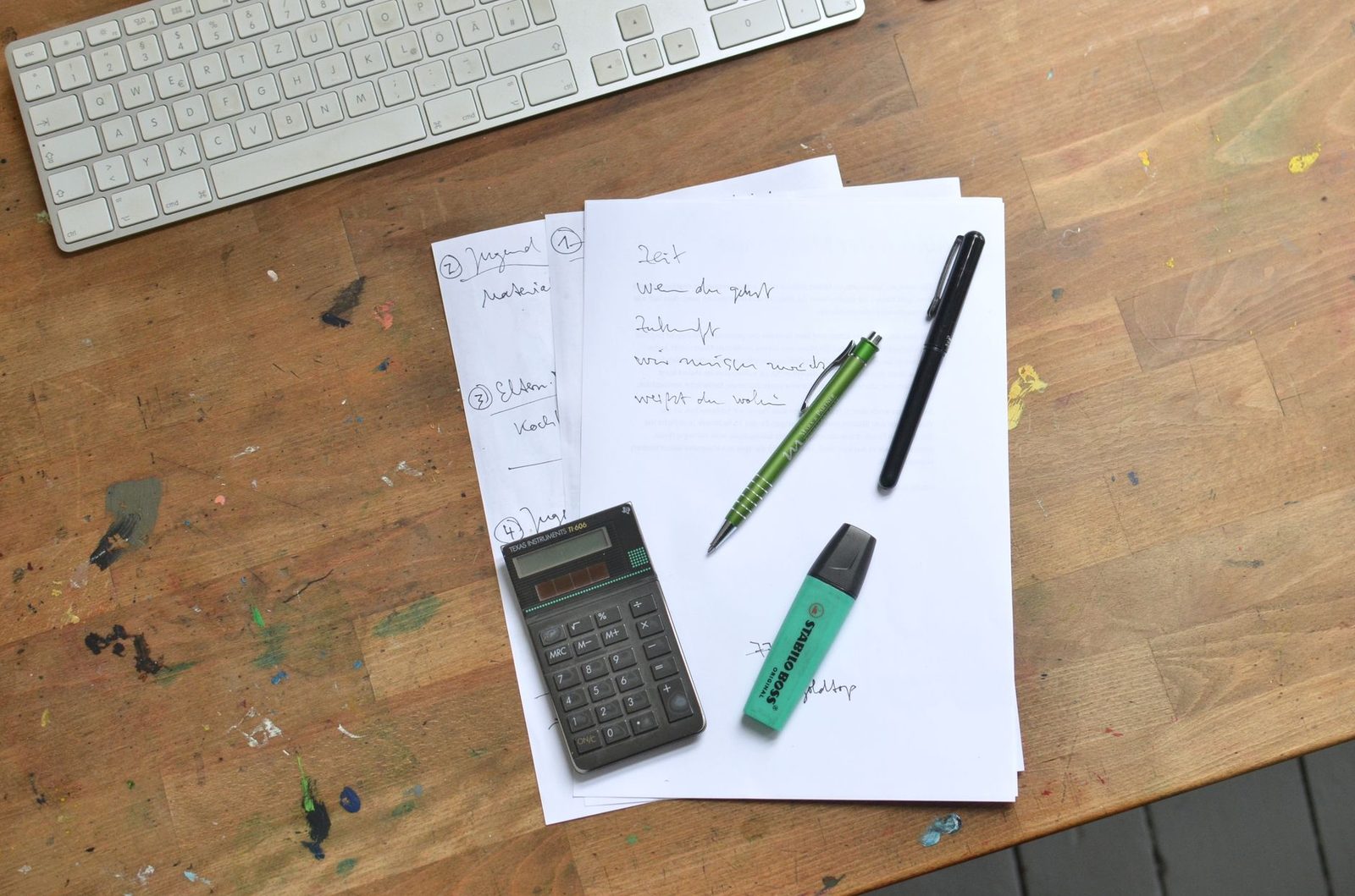 Top view of paper sheets with notes placed with pens with calculator and marker near keyboard on dirty table