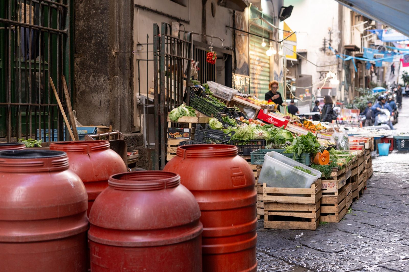 A lively urban market street scene with fresh produce and red barrels displaying local culture.