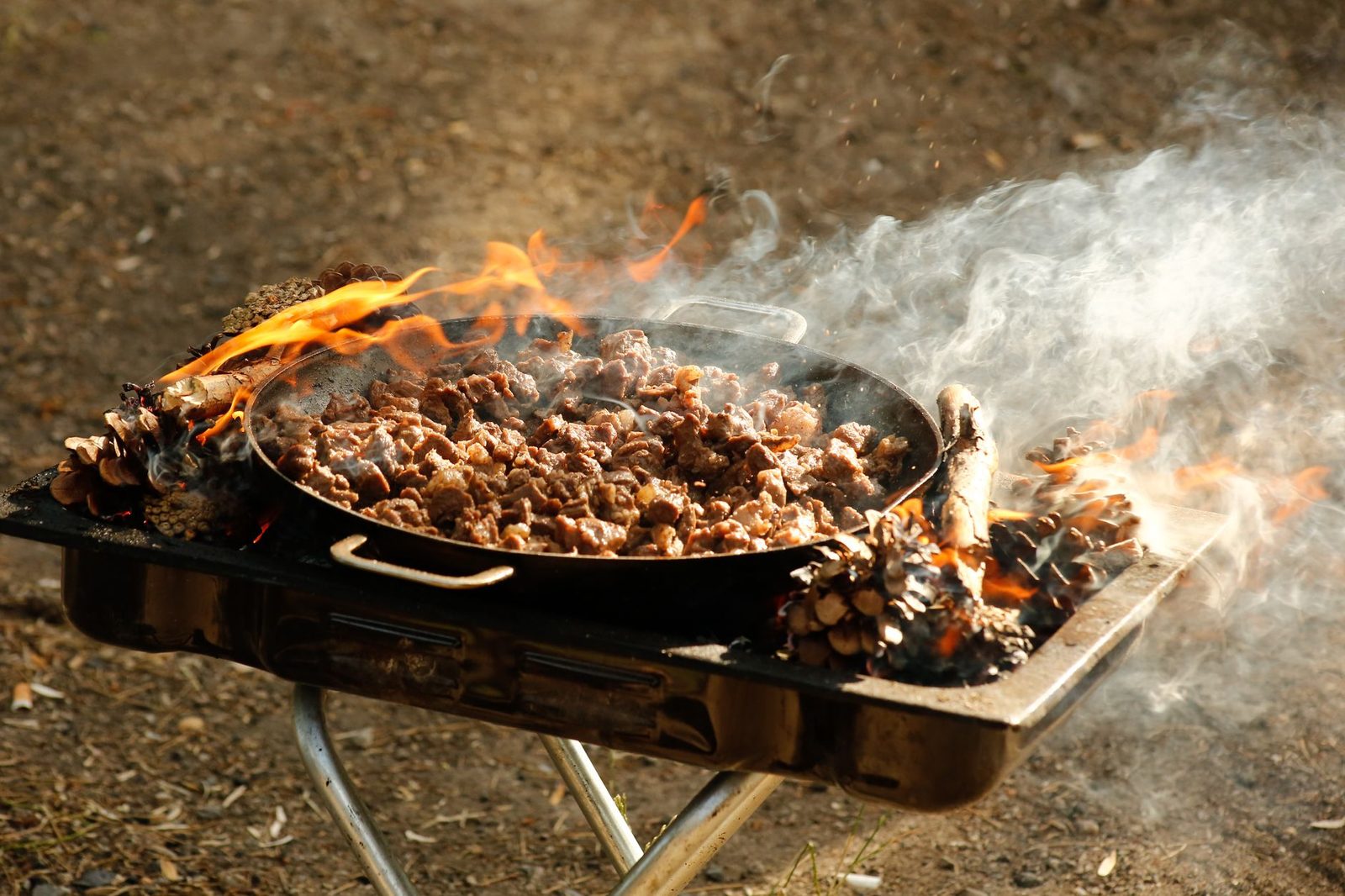 Close-up of sizzling meat cooking on an outdoor barbecue grill surrounded by flames and smoke.