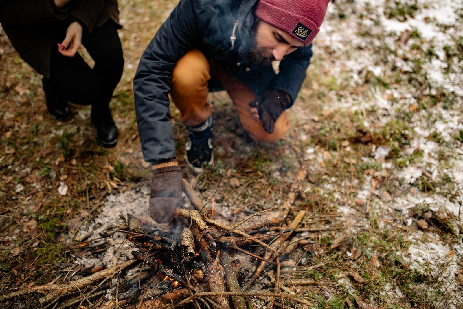 A man tending to a small campfire amidst a light snow-covered ground outdoors.