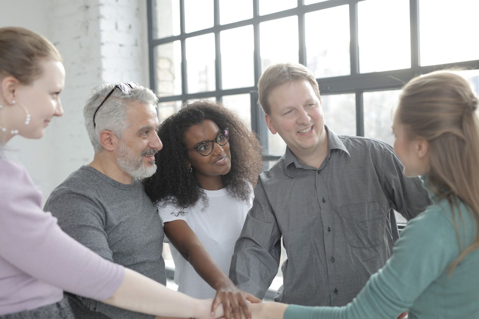 Happy multiethnic partners in casual clothes uniting hands while standing near fenced window in sunlight and looking at each other