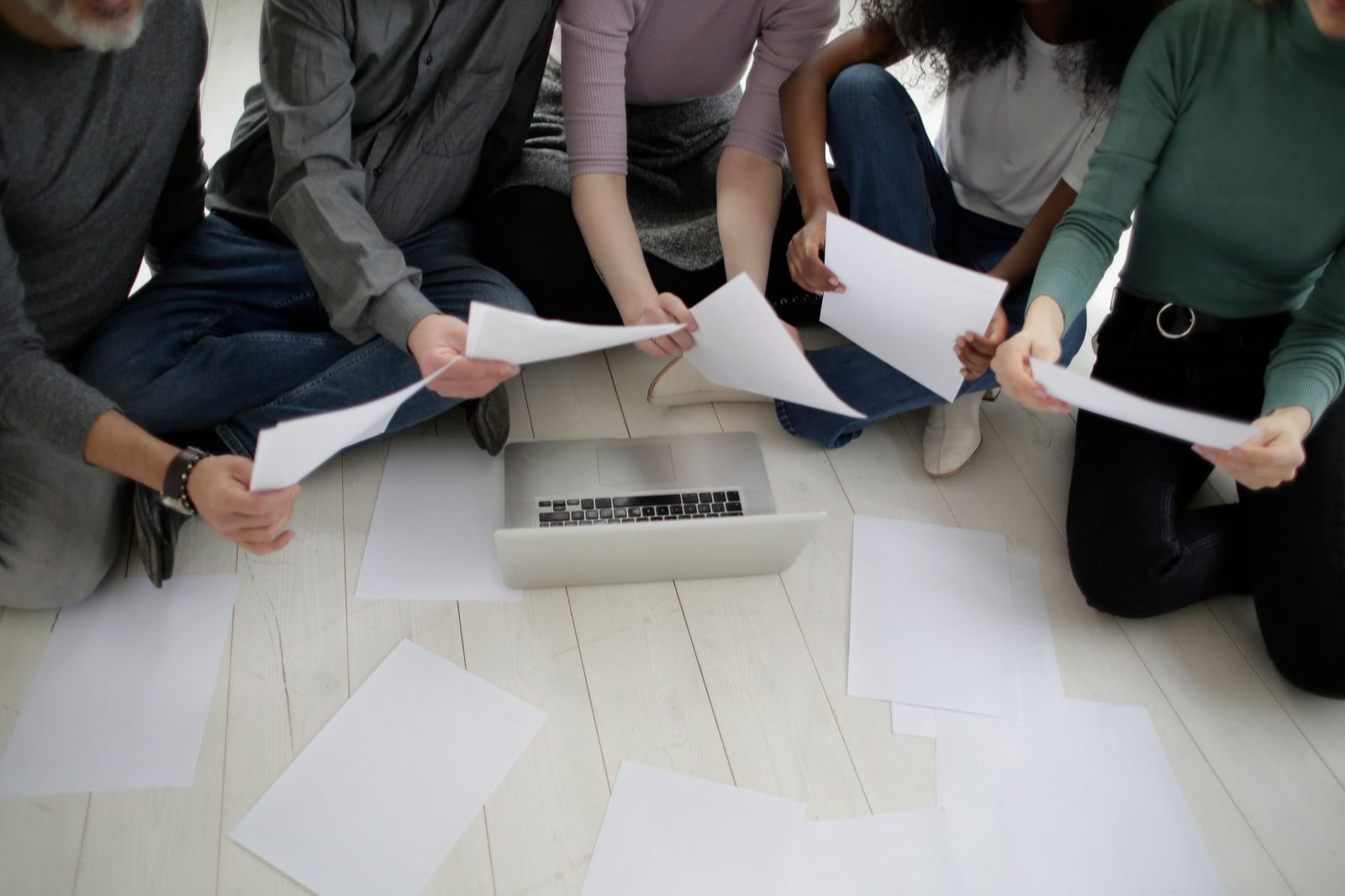 Crop faceless diverse coworkers in casual wear collecting assorted empty white papers sitting on floor in front of netbook in room