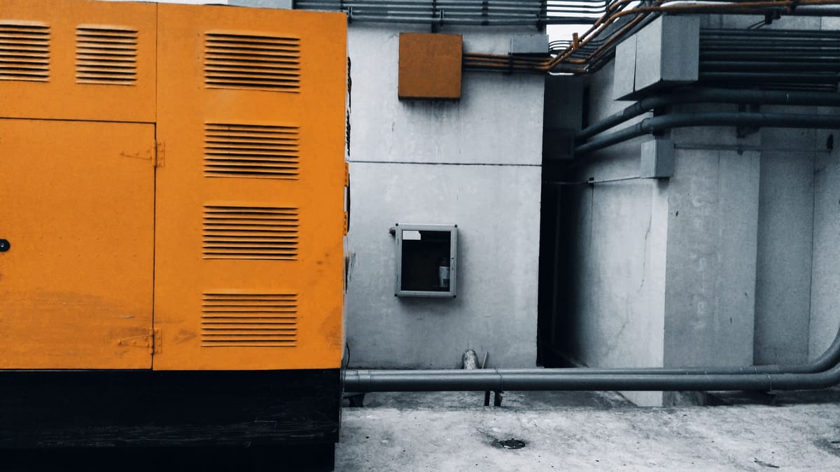 Outdoor view of an industrial generator with visible piping and wiring against a concrete wall.