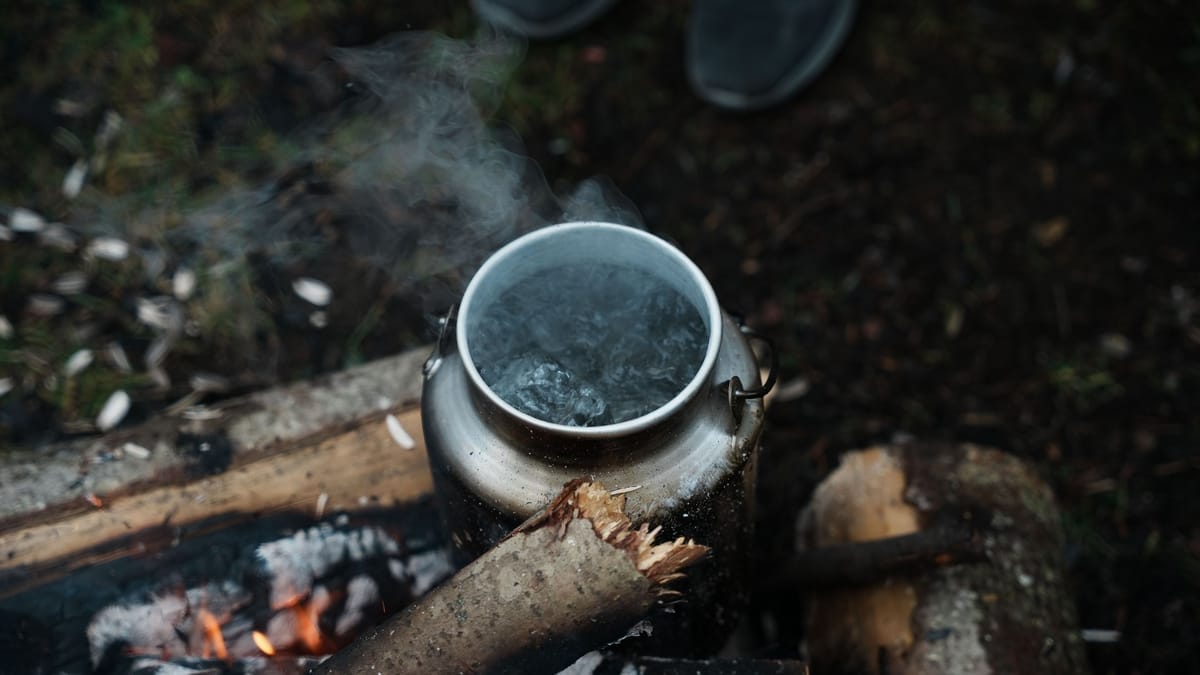 A silver kettle steaming as it boils water over a rustic campfire in the woods. Outdoor camping scene.