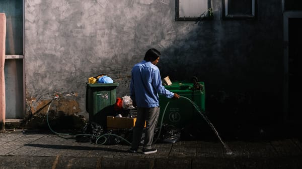 A person uses a hose to clean the street in a Vietnamese urban setting. Daylight scene with visible garbage bins.