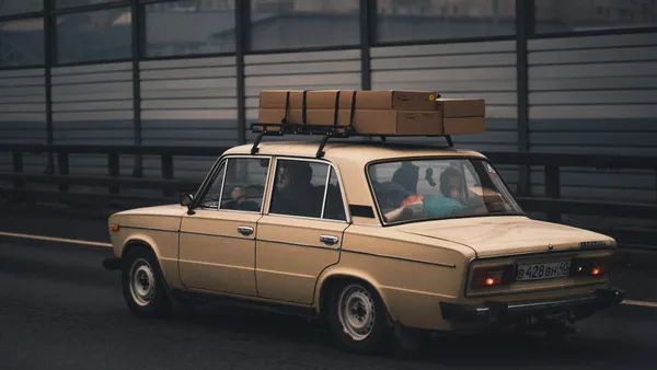Classic beige sedan carrying luggage, driving on highway with city in background.