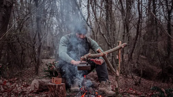 A man prepares food over a campfire in a dense forest, highlighting the essence of wilderness survival.