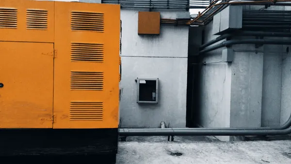 Outdoor view of an industrial generator with visible piping and wiring against a concrete wall.