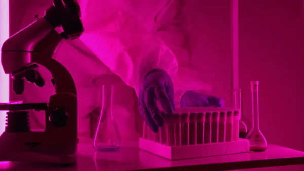 Scientist in a lab coat handling test tubes under pink lighting, using a microscope.