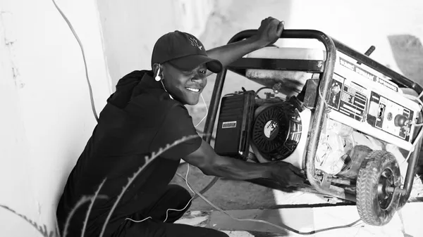 A cheerful young man wearing a cap and earphones operating a portable generator outside.