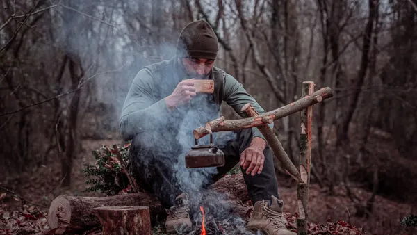 An adult man enjoys coffee by a campfire in a serene forest setting.