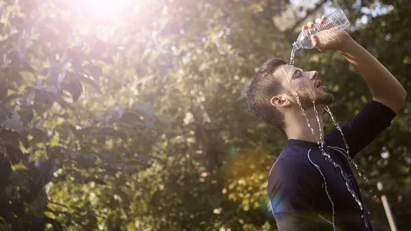 A man pours water over his head to cool off on a sunny day in a leafy outdoor setting.