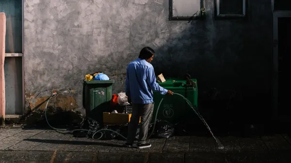 A person uses a hose to clean the street in a Vietnamese urban setting. Daylight scene with visible garbage bins.