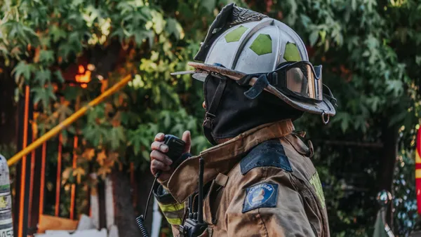 Firefighter in full protective gear using a radio outdoors.