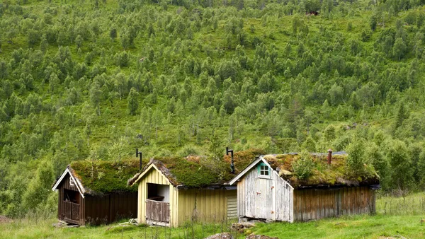 Rustic wooden cabins with moss-covered roofs nestled in lush green hills and forest.