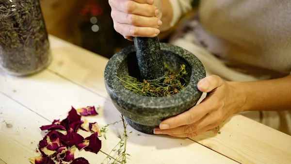 A person grinding herbal ingredients in a stone mortar and pestle with dried petals nearby.
