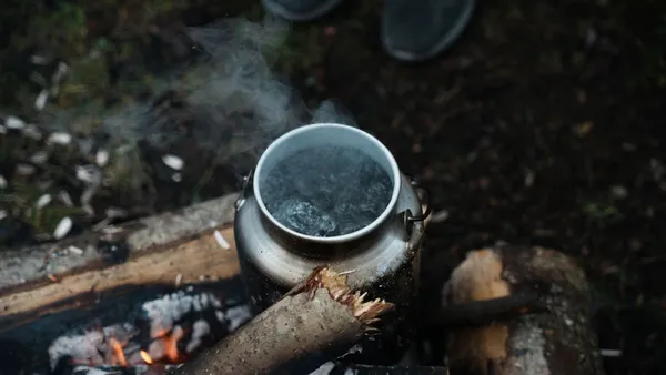 A silver kettle steaming as it boils water over a rustic campfire in the woods. Outdoor camping scene.