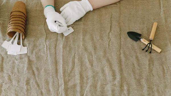 Person preparing gardening tools and labels on burlap surface, wearing gloves.