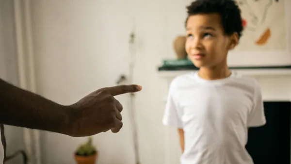 Crop unrecognizable strict black man scolding and disciplining guilty attentive little son standing with hands behind back and looking up