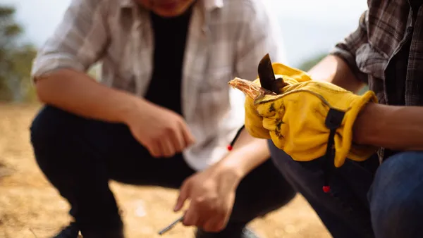 Two adults practicing bushcraft activities outdoors with gloves and knife skills.